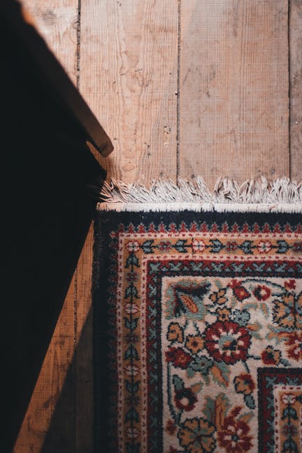 A close-up view of a traditional patterned rug with floral motifs in shades of red, blue, beige, and green, situated on a wooden floor with visible grain and knots. The rug features a fringed edge alo